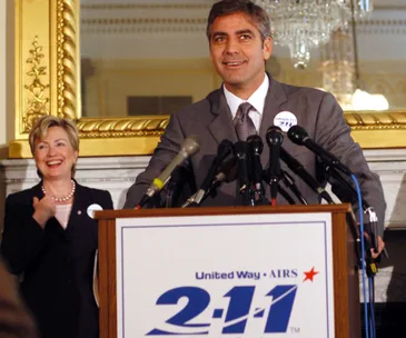Alt text: "Two people stand at a podium with microphones in a formal setting, one speaking and the other smiling."