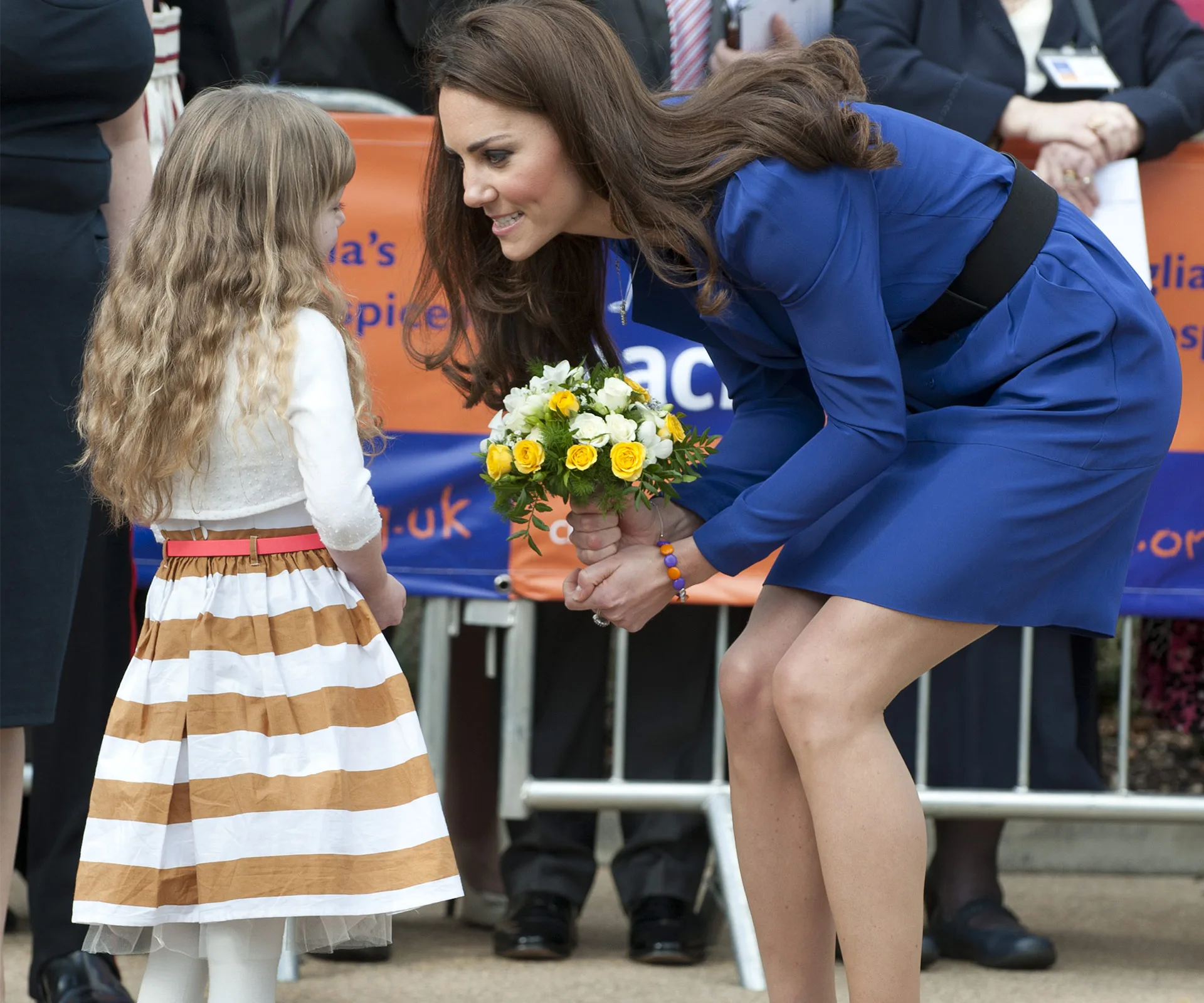 A woman in a blue dress is crouching to talk to a young girl, holding a bouquet of yellow and white flowers.