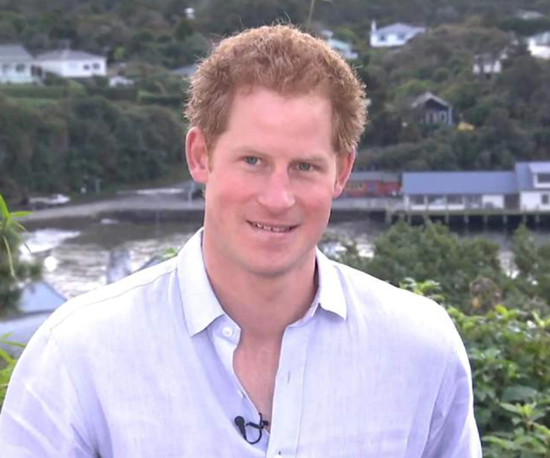 A man in a light blue shirt stands outdoors, with a river and houses in the background.