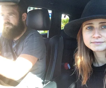 A man and woman sitting in a car, with the woman wearing a black hat and looking at the camera.