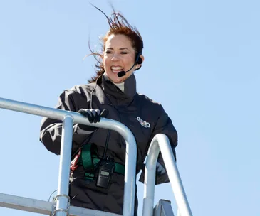 A person smiling while climbing a bridge, wearing a headset and safety gear, with wind-blown hair against a clear sky.