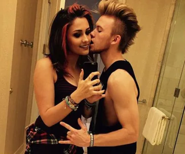 Two people taking a mirror selfie, one kissing the other's cheek, in a bathroom setup.