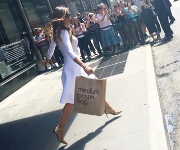 Woman in white dress with shopping bag, high heels, walking on a sunny sidewalk near a crowd taking photos.