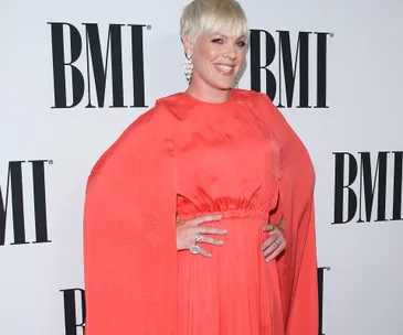 Singer in a coral dress smiling at a BMI event, hands on hips against a backdrop with BMI logos.