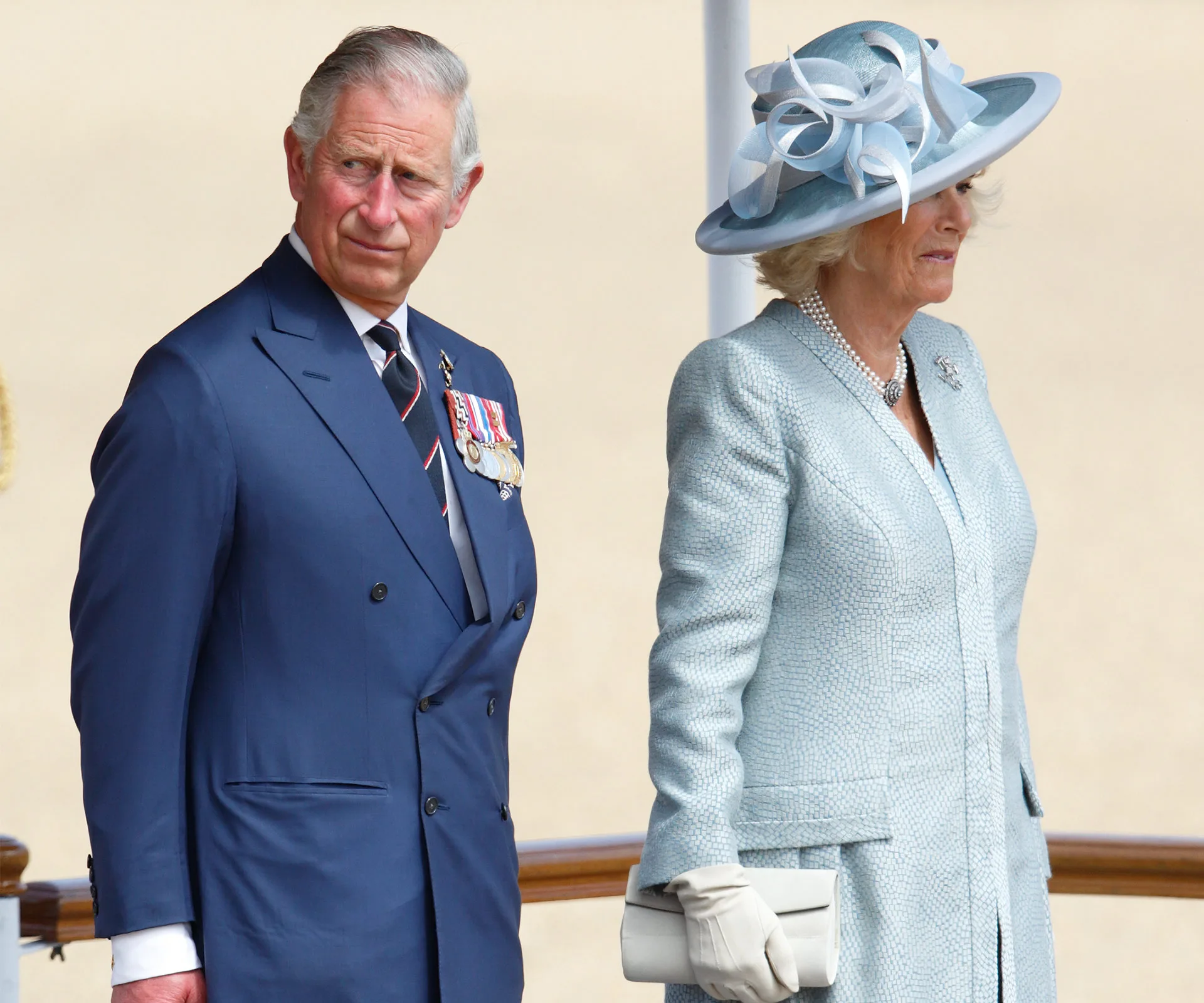 Royal couple in formal attire, with medals and a blue suit, stand together at an outdoor event.