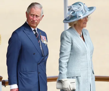 Royal couple in formal attire, with medals and a blue suit, stand together at an outdoor event.