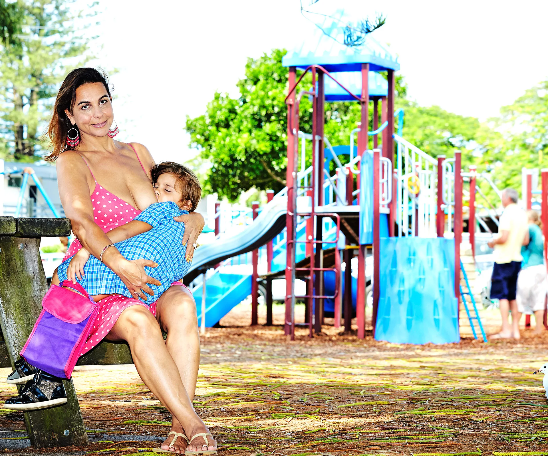 A woman in a pink dress is breastfeeding a child on a bench at a playground.