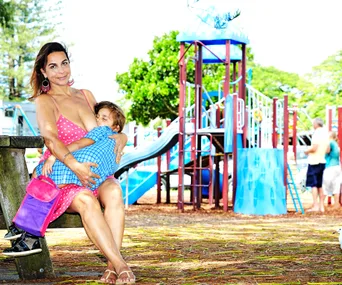 A woman in a pink dress is breastfeeding a child on a bench at a playground.