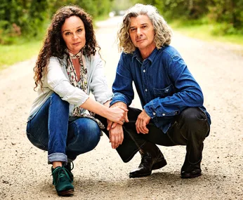 Two people crouching on a dirt road, surrounded by greenery, holding hands and looking at the camera.