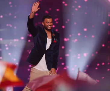 Guy Sebastian waving on stage during Eurovision performance, surrounded by pink lights and flags.
