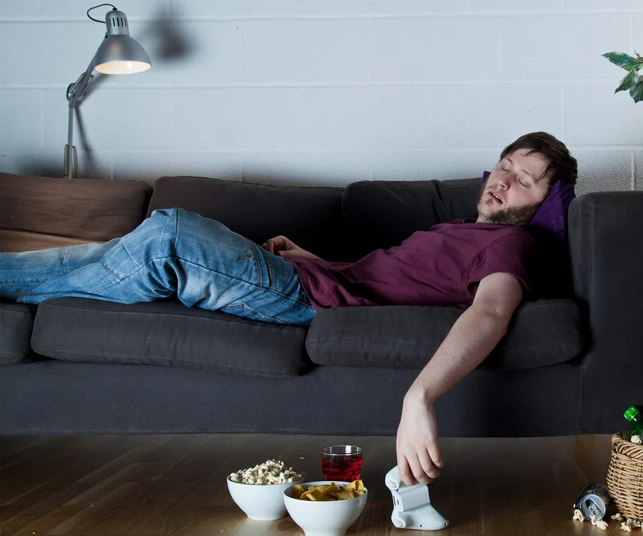 Man sleeping on a couch, surrounded by snacks and a drink, holding a video game controller.
