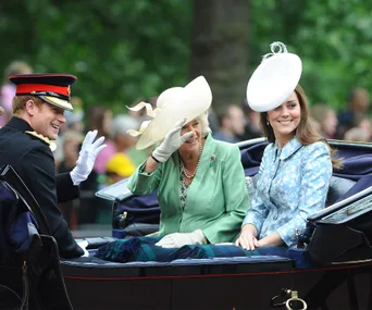 Three people in formal attire ride in a carriage, smiling and waving, surrounded by greenery.