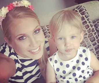 Smiling woman with flower headband and striped top, sitting with a young child in a polka dot dress on a patterned couch.