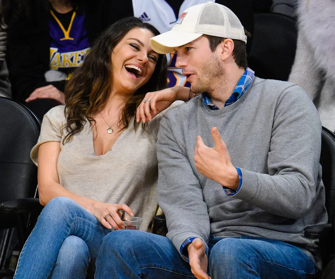A couple laughing and enjoying a basketball game, sitting in the stands, with casual attire and a Lakers fan in the background.