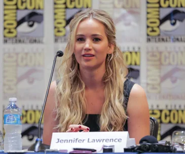 Jennifer Lawrence speaking at a Comic-Con panel, seated with name placard and microphone.