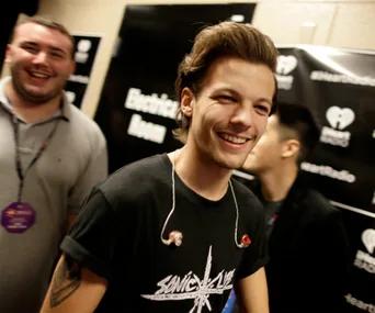 "Man smiling backstage at an event with others in the background, wearing a black t-shirt and an in-ear monitor."