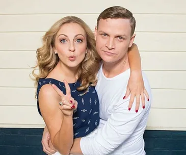 Pair posing playfully, woman making a peace sign and man embracing her, in front of a light-colored wall.