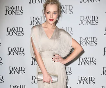 A person in an elegant dress poses confidently at a David Jones event, holding a matching clutch against a branded backdrop.