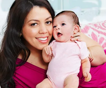Smiling woman holding a baby wearing pink, both looking towards the camera.