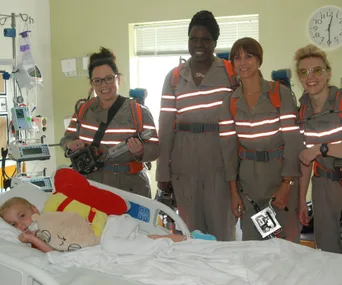 Ghostbusters cast in uniform visit a child in a hospital bed, bringing smiles and joy with proton packs and gear.
