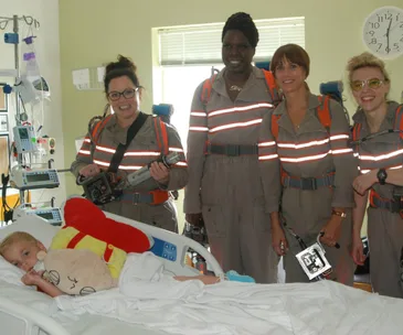 Ghostbusters cast in uniform visit a child in a hospital bed, bringing smiles and joy with proton packs and gear.
