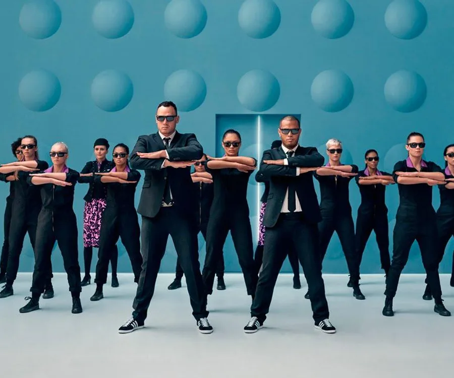 Men in black suits and sunglasses leading a group in black uniforms against a blue backdrop with circular patterns.