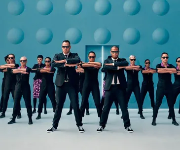 Men in black suits and sunglasses leading a group in black uniforms against a blue backdrop with circular patterns.
