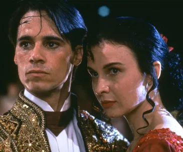 Two dancers dressed in ornate costumes, looking serious and intense, under dim lighting.