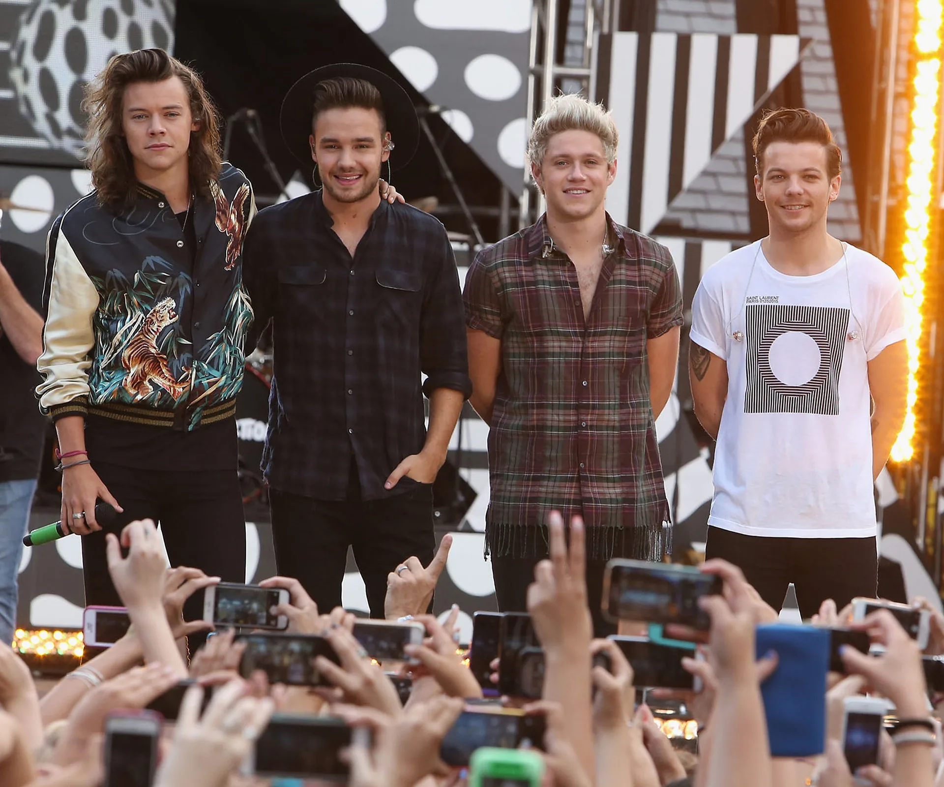 One Direction band members standing on stage with fans raising phones for photos.