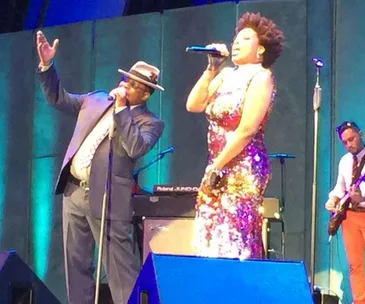 Singer in a suit and hat performing on stage with a female singer in a sequined dress, accompanied by a guitarist.