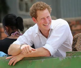 A man smiles, leaning on a green surface, wearing a white shirt in an outdoor setting, with blurred background figures.