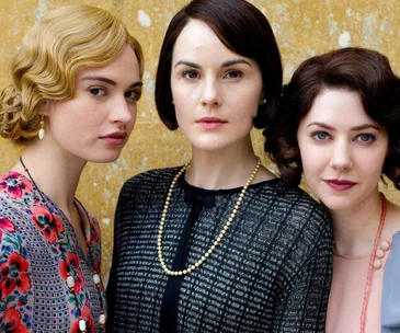 Three women in period attire from "Downton Abbey" stand close, showcasing distinct 1920s hairstyles against a textured background.