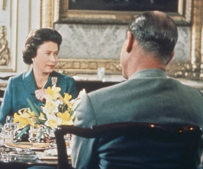 Dining scene with a woman in a blue dress and a man in a gray suit, a floral centerpiece visible between them.