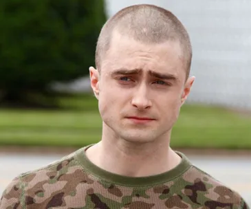 Actor with a shaved head wearing a camo shirt, standing outside with a blurred background.