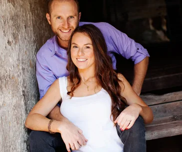 Couple smiling warmly, the woman in a white top seated with the man in a purple shirt embracing her from behind.