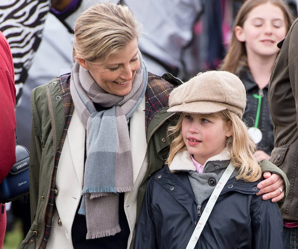 A woman in a green jacket and scarf smiles at a young girl in a flat cap and raincoat outdoors.