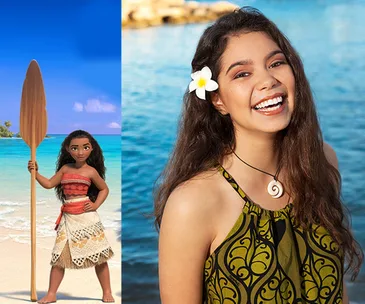 "Moana character holding a paddle on the beach next to actress with a flower in her hair, smiling by the ocean."