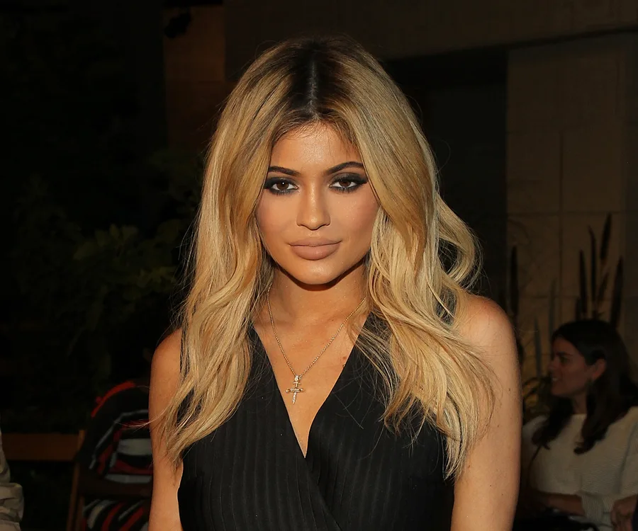 ```plaintext
Kylie Jenner in a black dress with blonde hair, wearing cross necklace.
```