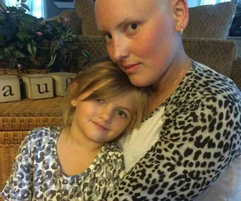 A young girl cuddles with her bald mother, who is smiling, both wearing matching leopard print tops.