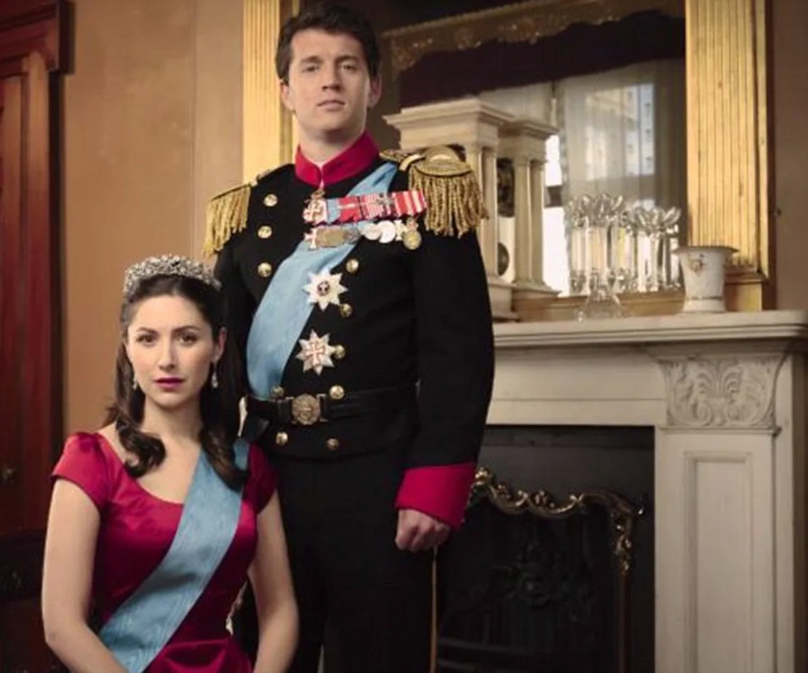 Royal couple in formal attire, with the woman seated wearing a tiara and the man standing in a uniform, in a regal room.