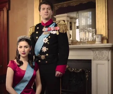 Royal couple in formal attire, with the woman seated wearing a tiara and the man standing in a uniform, in a regal room.