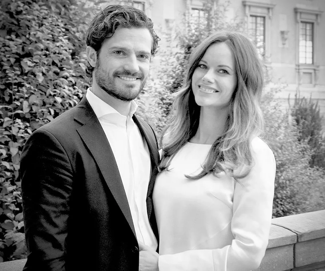 A smiling couple poses together, outdoors, dressed in formal attire.