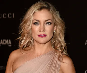 Kate Hudson at an event, wearing a one-shoulder dress, with blonde hair and bright lipstick, on a dark background.