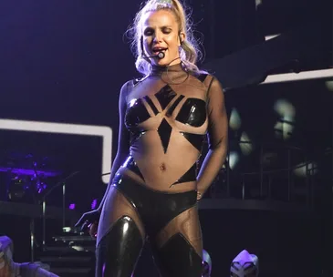 Singer performing on stage in a sheer black geometric outfit, smiling with a headset microphone.