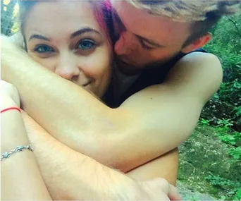 Young couple shares a sweet embrace outdoors, smiling, with a forest backdrop.