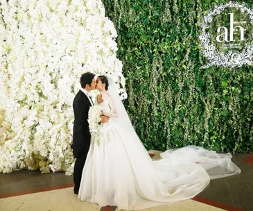 A bride and groom kiss in front of a floral wall, with the bride in a white gown holding a bouquet.