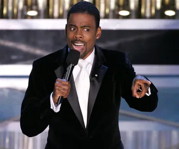 Chris Rock hosting the 2016 Oscars, wearing a tuxedo and holding a microphone, gesturing animatedly.