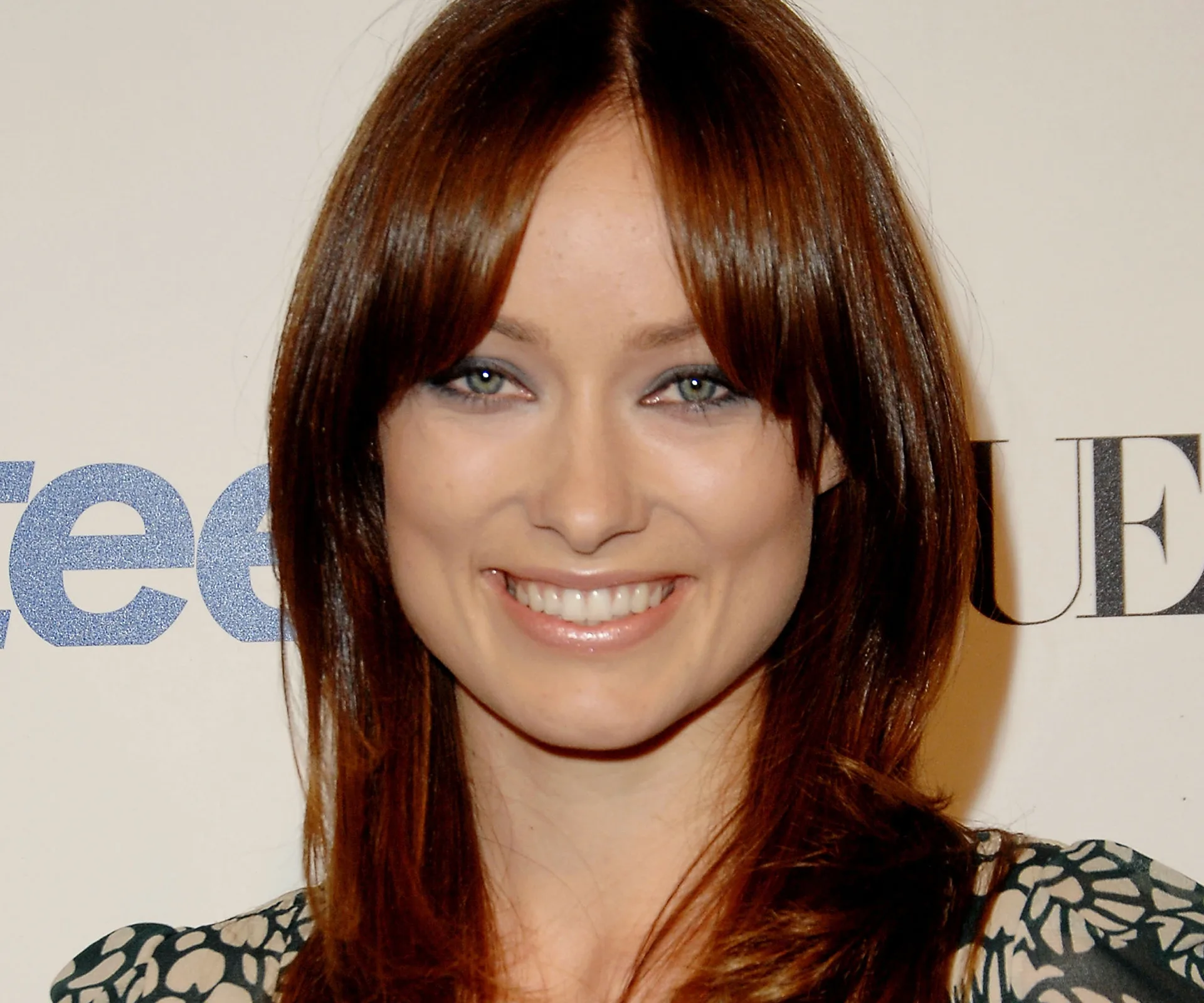 Smiling woman with long brown hair at an event, light floral-patterned dress, standing against a backdrop with logos.