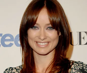 Smiling woman with long brown hair at an event, light floral-patterned dress, standing against a backdrop with logos.