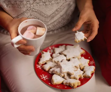 Person holding hot cocoa with marshmallows and a plate of star-shaped cookies in a cozy setting.